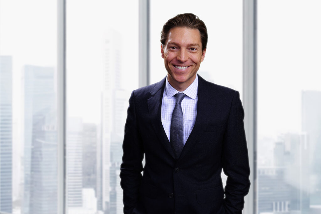 Man in dark suit, tie stands in front of windows with city skyline in background