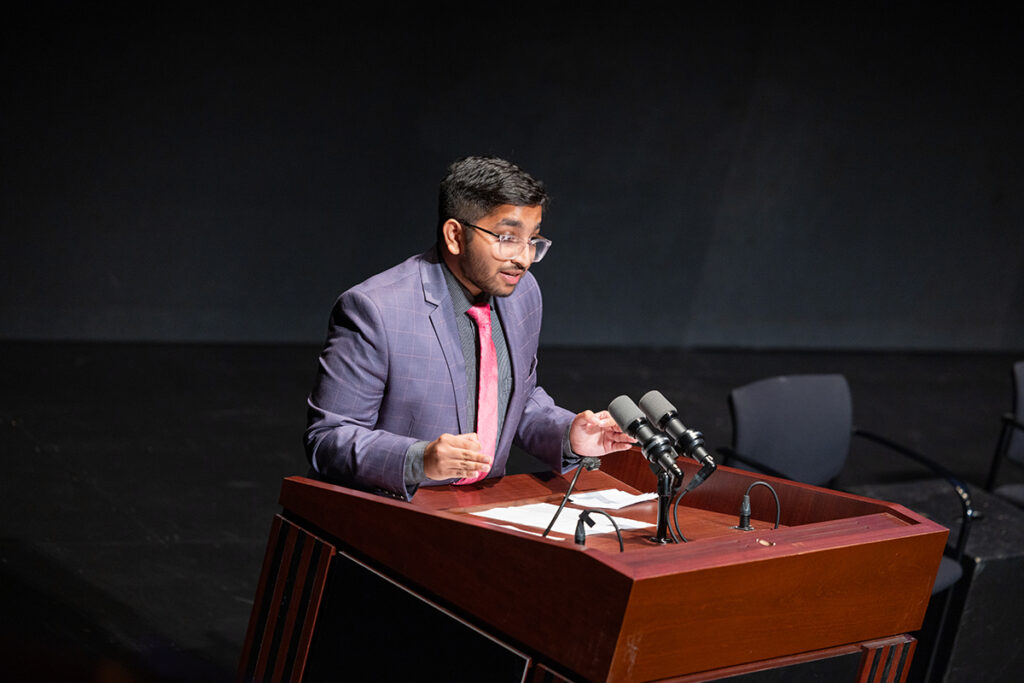 Man with short black hair wearing purple jacket, pink tie, clear-framed glasses, speaks at podium