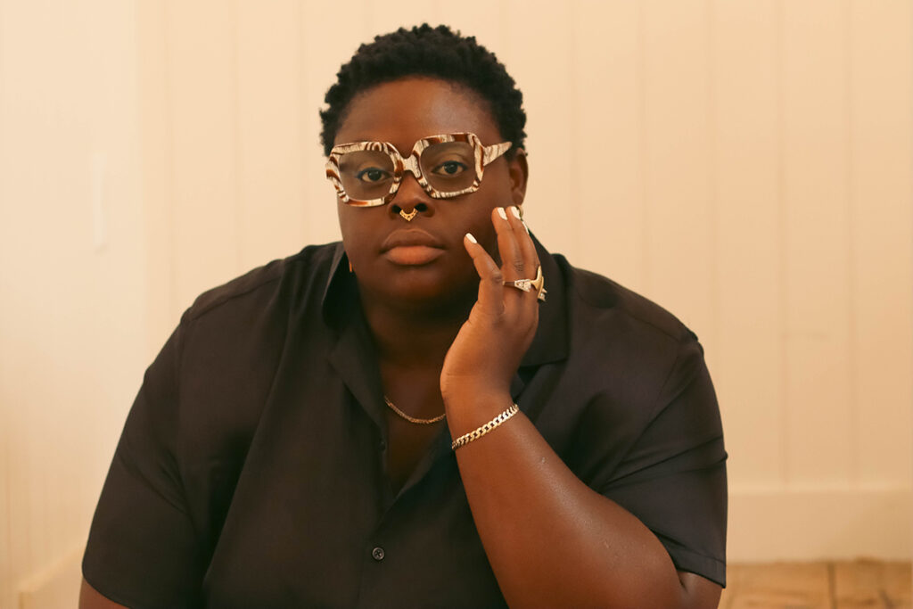 Black woman with short hair, thick tortoiseshell framed glasses, wearing a black shirt holds hand to her face