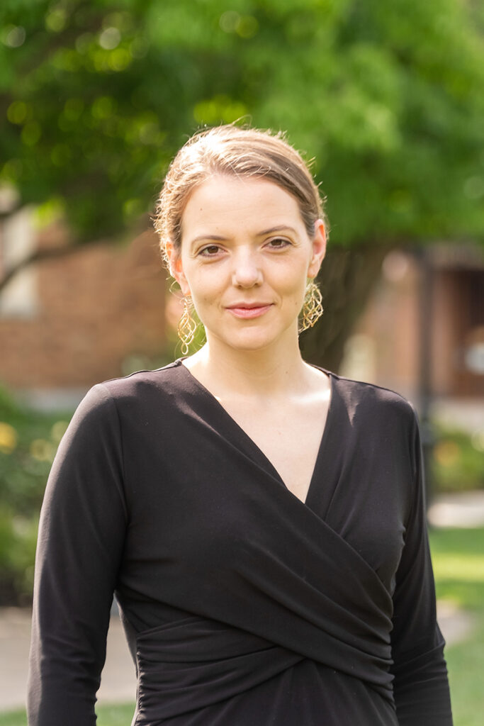 Woman with light brown hair, wearing black wrap dress and dangly gold earrings stands in front of tree and building