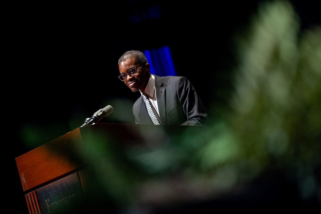 Man with greying hair, cropped close, wearing glasses, dark suit, blue and yellow plaid tie speaks at podium