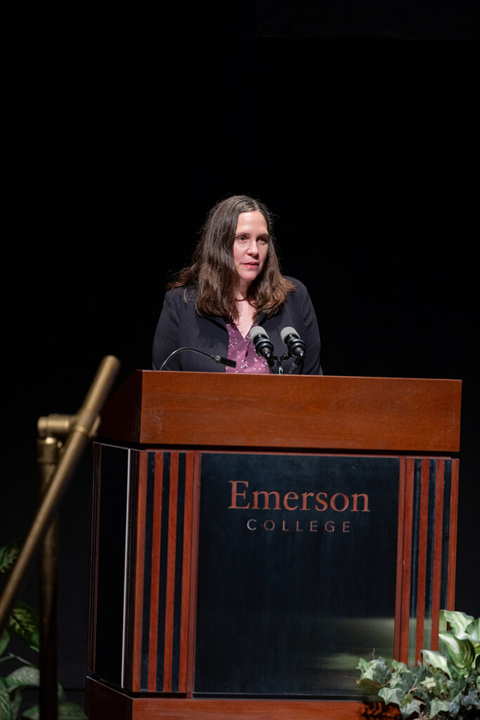 Woman with long brown hair, wearing a purple shirt and dark jacket, speaks at podium surrounded by plants