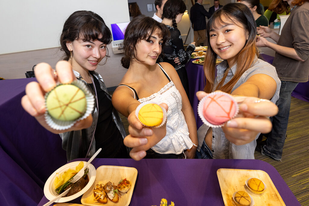 Three young women stand behind table covered with food, holding macarons toward the camera