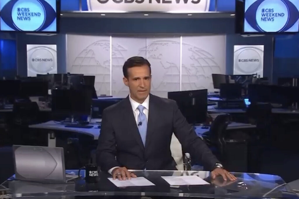 man in dark suit sits behind anchor desk with CBS Weekend News set behind him