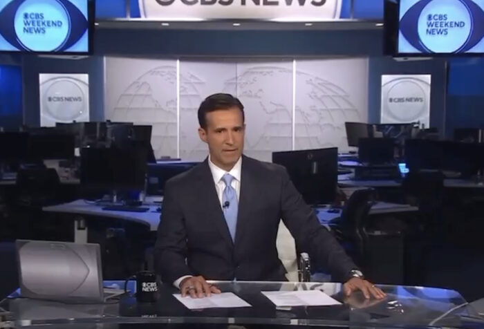 man in dark suit sits behind anchor desk with CBS Weekend News set behind him