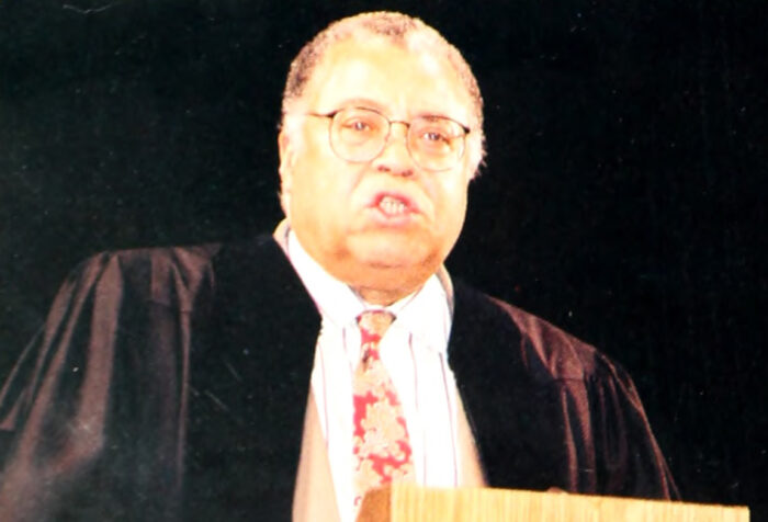 A man in a white shirt, tie, and black robe gives a speech