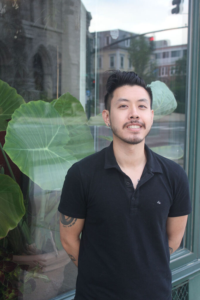 Person with short dark hair, short beard wearing black polo shirt stands in front of a window with plant behind it, city scape reflected in it