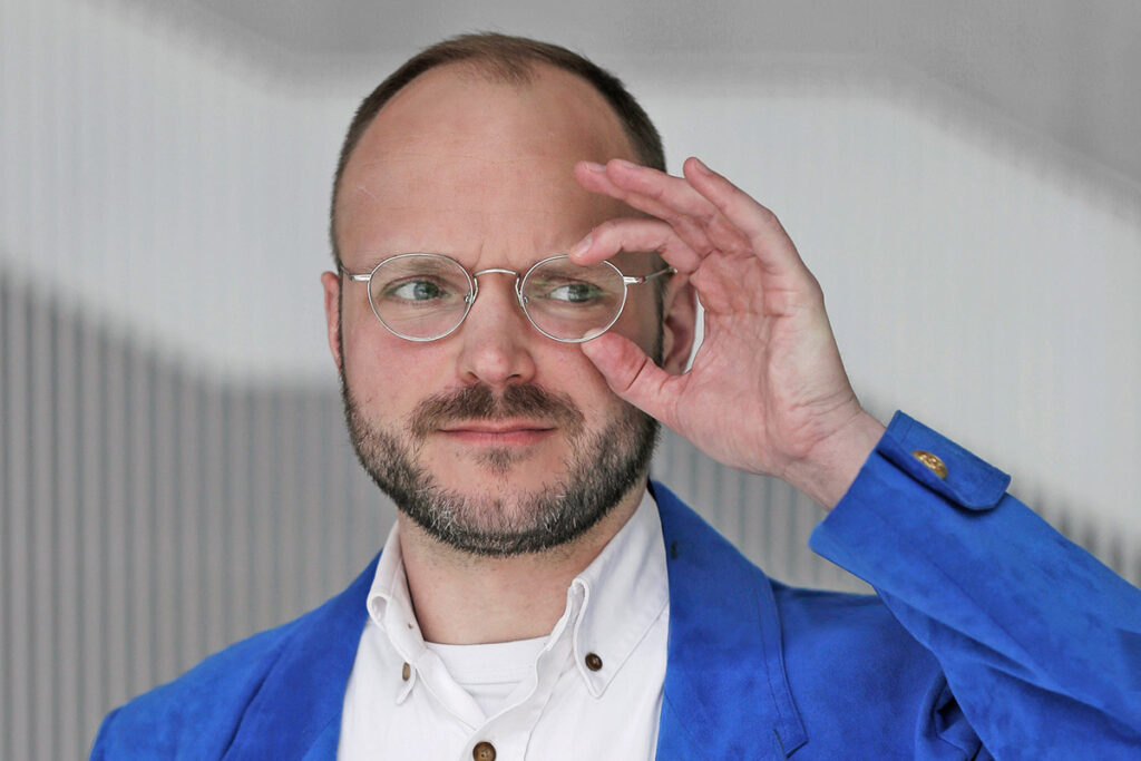 Man with close-cropped hair, beard, wearing white Oxford shirt and blue suede jacket touches a lens of his wire-framed glasses