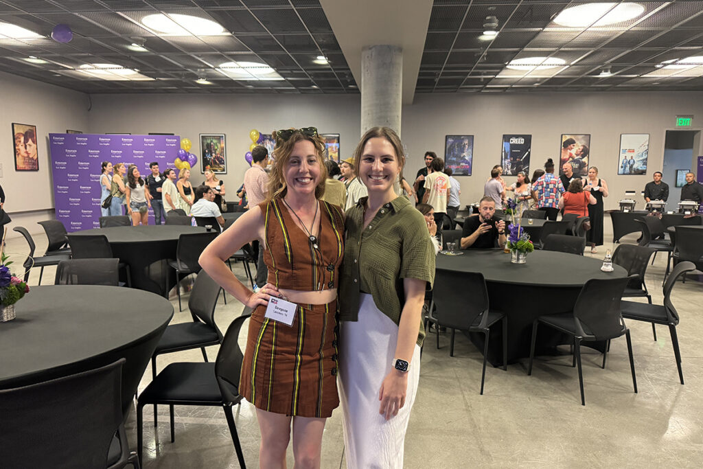 Two women, one wearing a brown and yellow striped skirt and top, and one in green shirt, white pants stand in a room with black tables set up