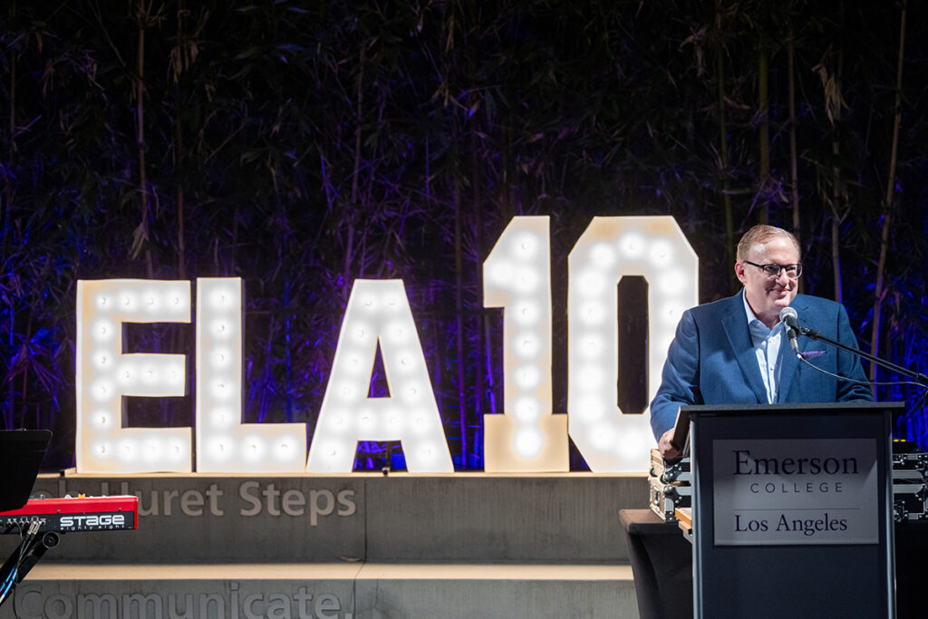 Man in blue suit, glasses, speaks at podium in front of sign of lights reading ELA 10