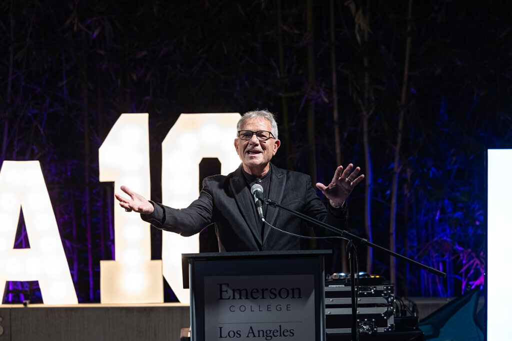 Man in black suit, glasses, speaks at podium with arms outstretched