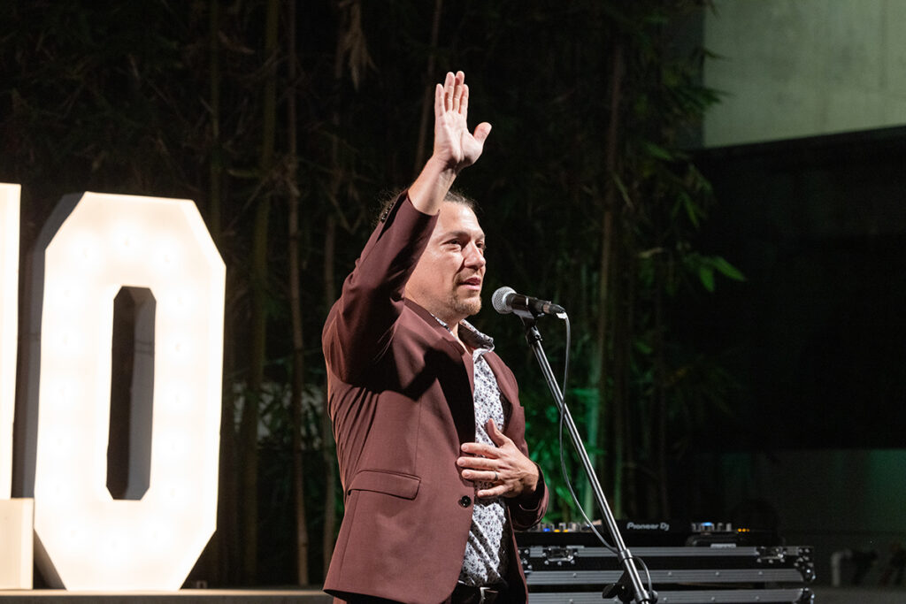 Man in print shirt, brown jacket stands before a microphone, raises hand