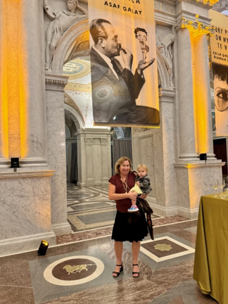 Woman in red top, black skirt, with medium length blonde hair, holds a toddler wearing a yellow dress in an ornate marble lobby