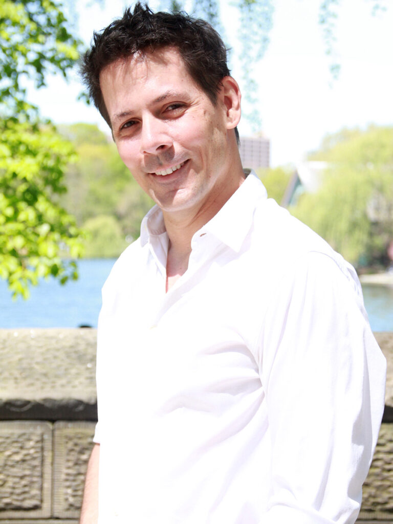 Man with short dark hair, wearing white button-down shirt stands on a bridge over a pond 