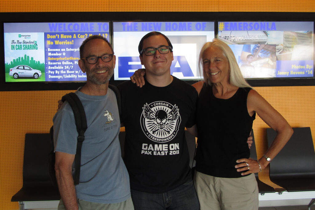 young man in glasses, black t-shirt flanked by man in blue t-shirt, backpack, and woman with long blonde hair wearing a black tank top