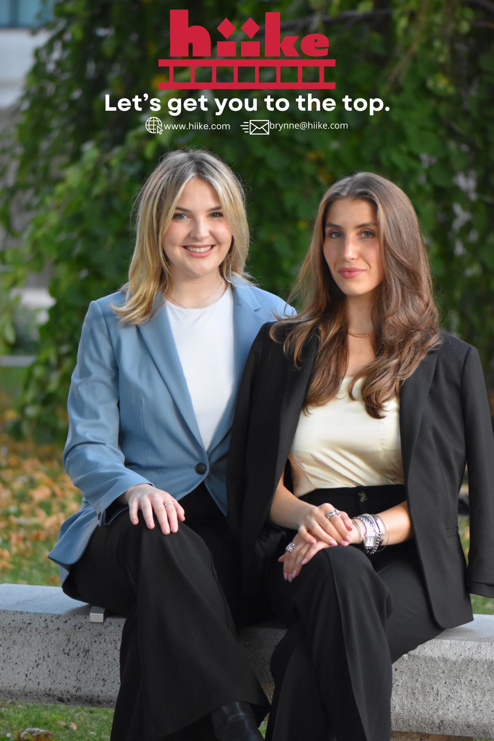 Two people wearing blazers pose for a photo, with a tree in the background.