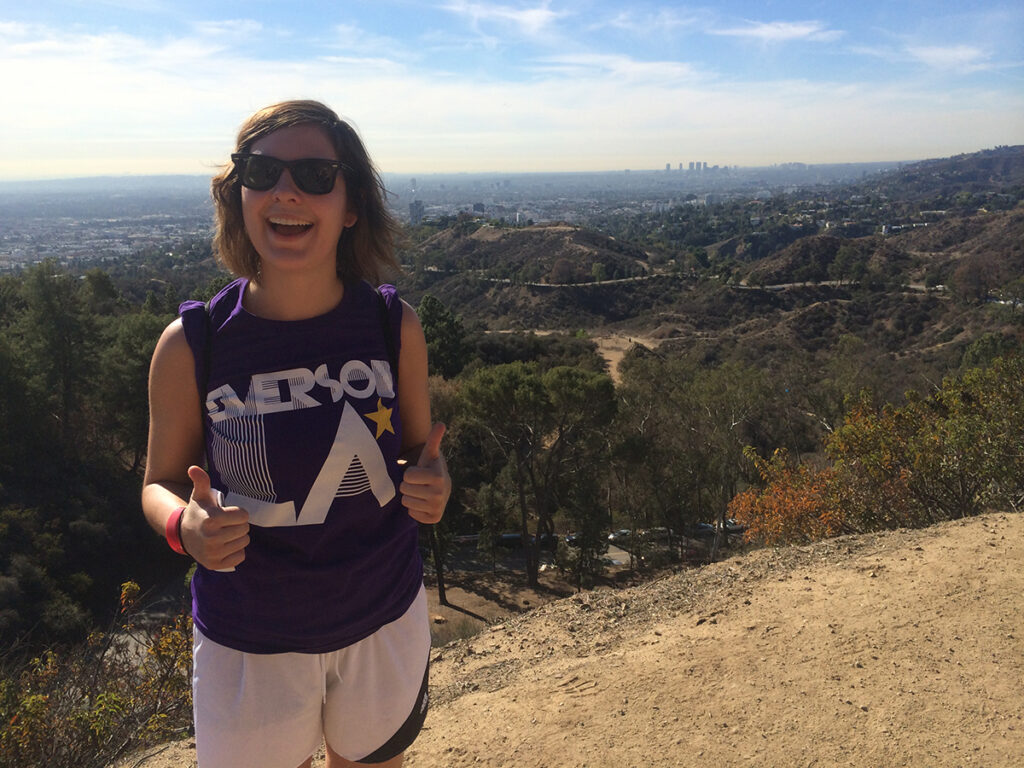 young woman in sunglasses, purple sleeveless Emerson LA t-shirt gives thumbs up with view of valley behind her