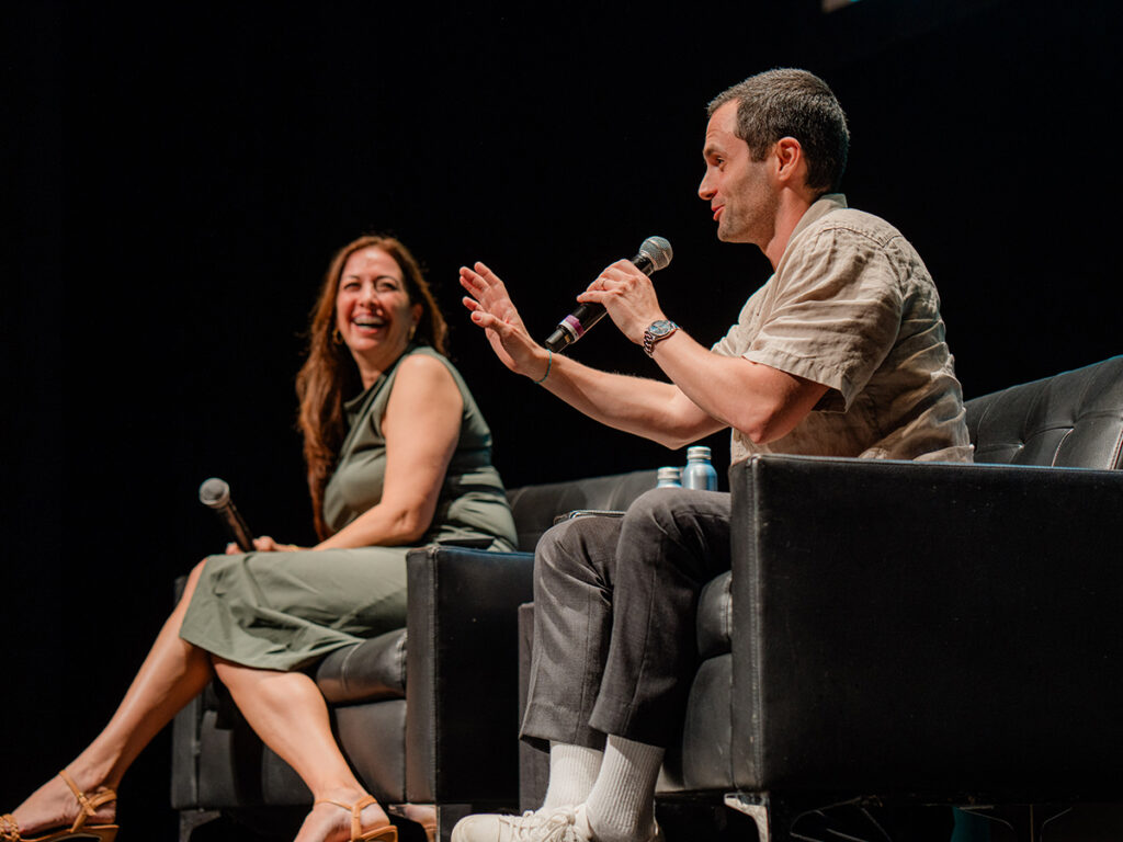 Woman in green dress, man in beige shirt speak into mics on stage