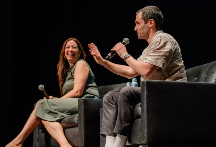 Woman in green dress, man in beige shirt speak into mics on stage