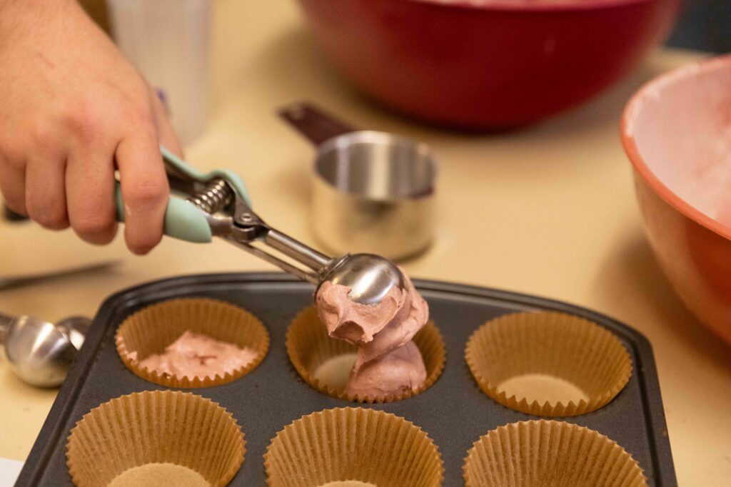 A scooper dropping blood red velvet cupcakes into a cupcake tin