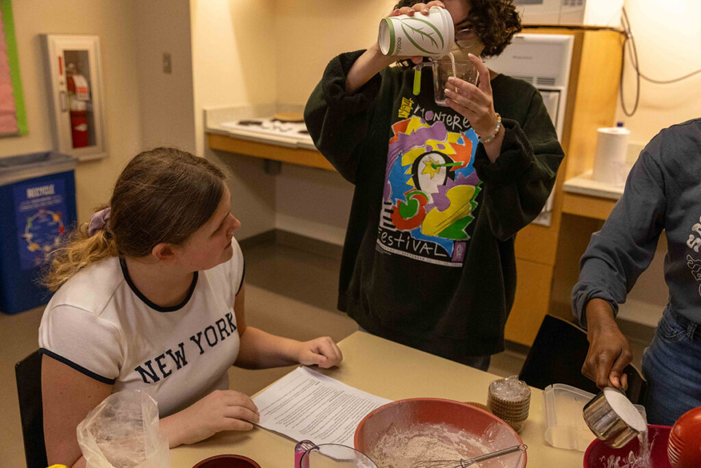 Student in measures pink batter from a paper cup into a measuring cup