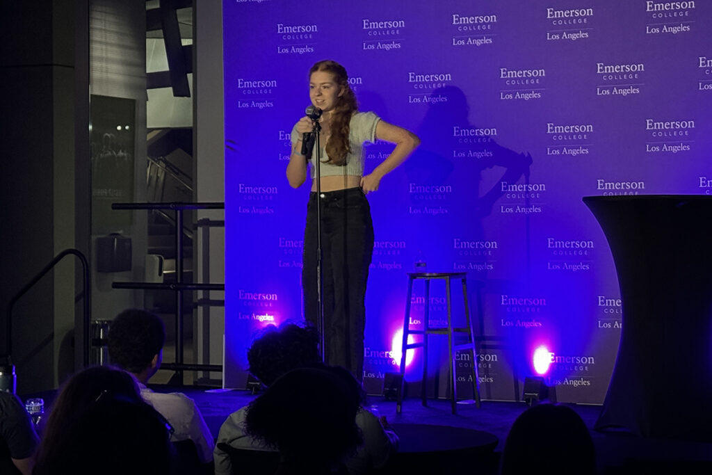 Girl with long red hair, wearing crop top and dark pants talks into a mic on a purple-hued stage