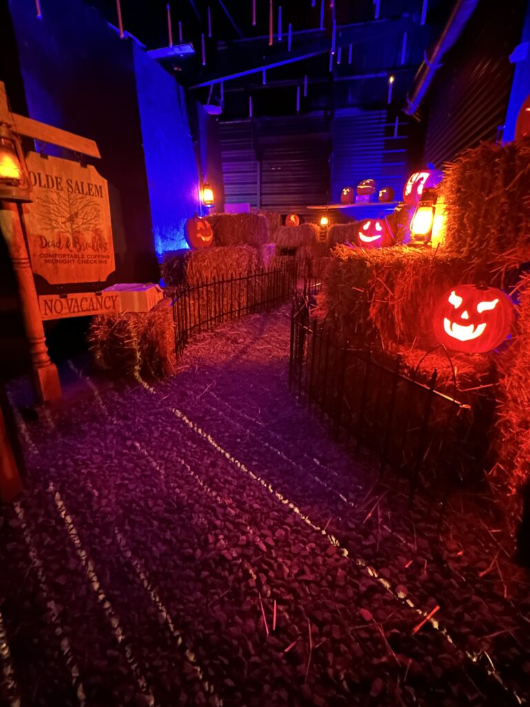 A dark room with lit jack o' lanterns on bales of hay, and a sign that says Salem.