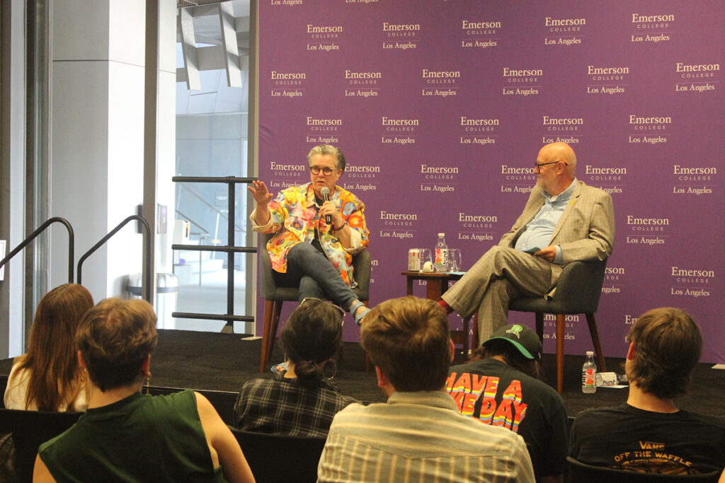 Rosie O'Donnell, wearing bright floral jacket, speaks into a mic as a man in a tan suit sits next to her and students in audience sit with back to camera