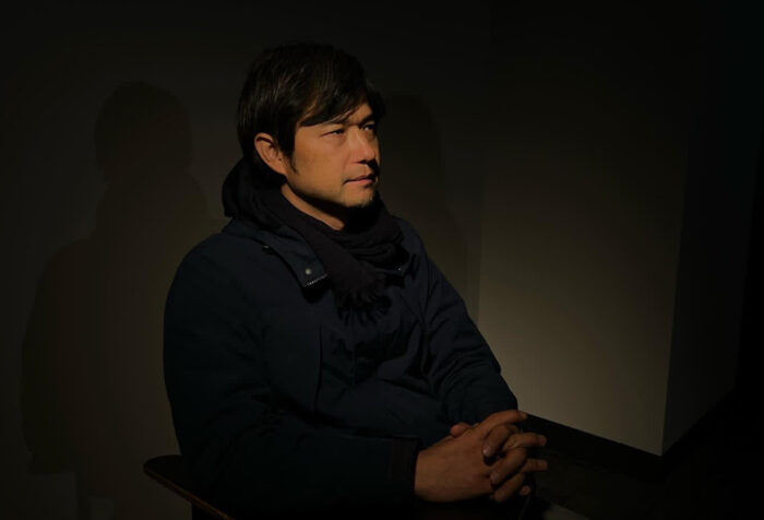 Man sits in profile wearing dark jacket, in low light