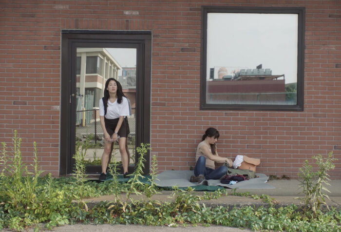 woman stands against glass door in brick building, another woman sits on the ground beneath window