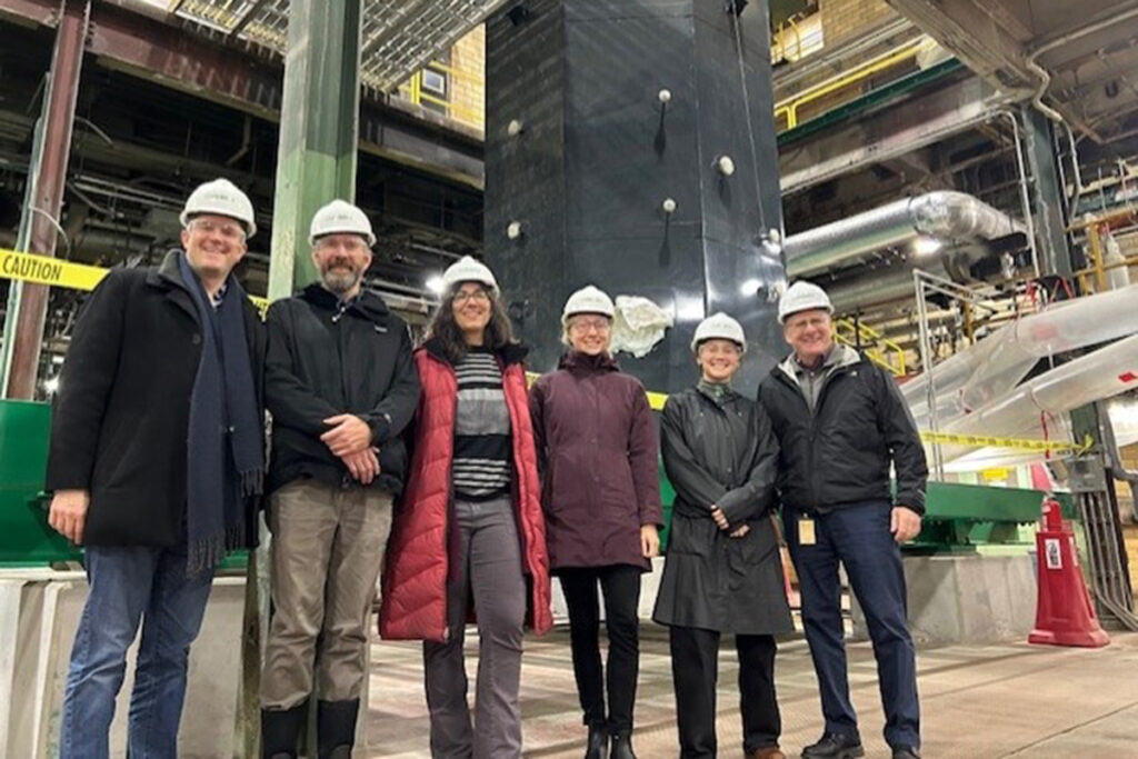 Six people wearing white hard hats stand in steam plant