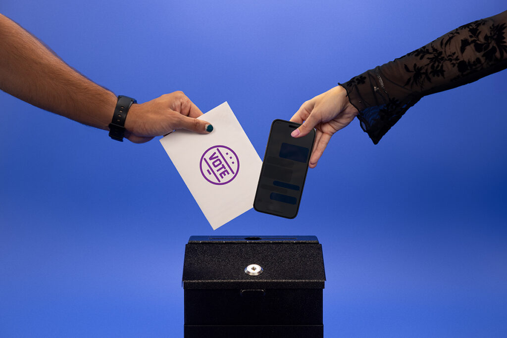 On hand places a phone into a voting box and another hand places an Emerson College Polling paper into the voting box