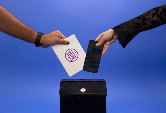 On hand places a phone into a voting box and another hand places an Emerson College Polling paper into the voting box