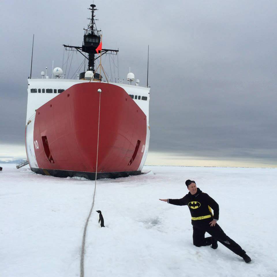 Justin Ramey in a Batman outfit on Antarctic ice by a little penguin with an ice breaker ship behind them with a rope coming from the boat