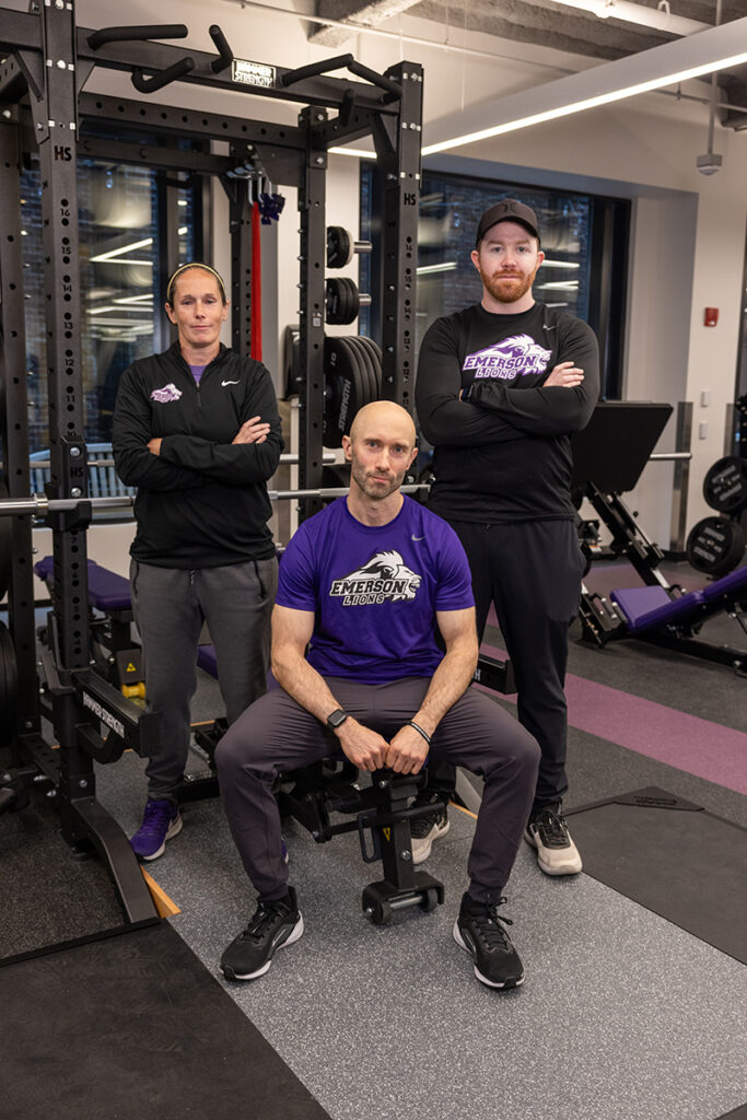 Man in purple Emerson t-shirt sits on weight bench, flanked by woman and man in black Emerson gear standing with arms crossed