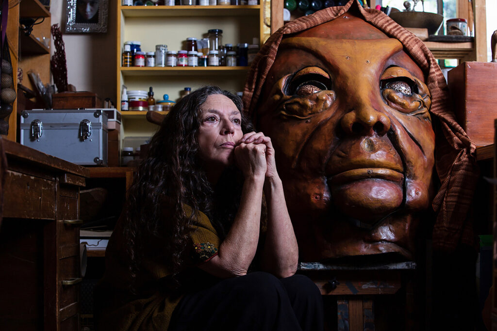 Woman sits with elbows on table as giant brown mask sits beside her