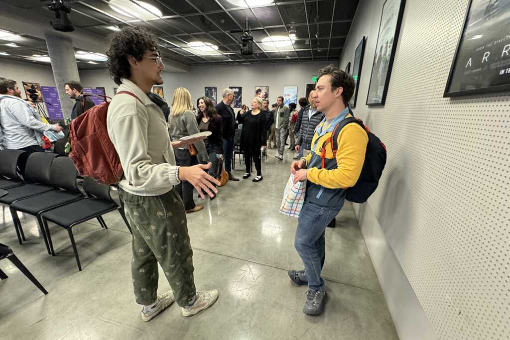 Man in green pants, backpack, talks to Brian Jordan Alvarez, also wearing a backpack, as crowd mills in background