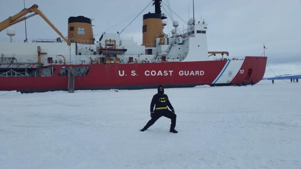 Justin Ramey in a Batman outfit on Antarctic ice with a U.S. Coast Guard ship behind him