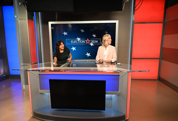 Women in professional clothing sit behind anchor desk in studio