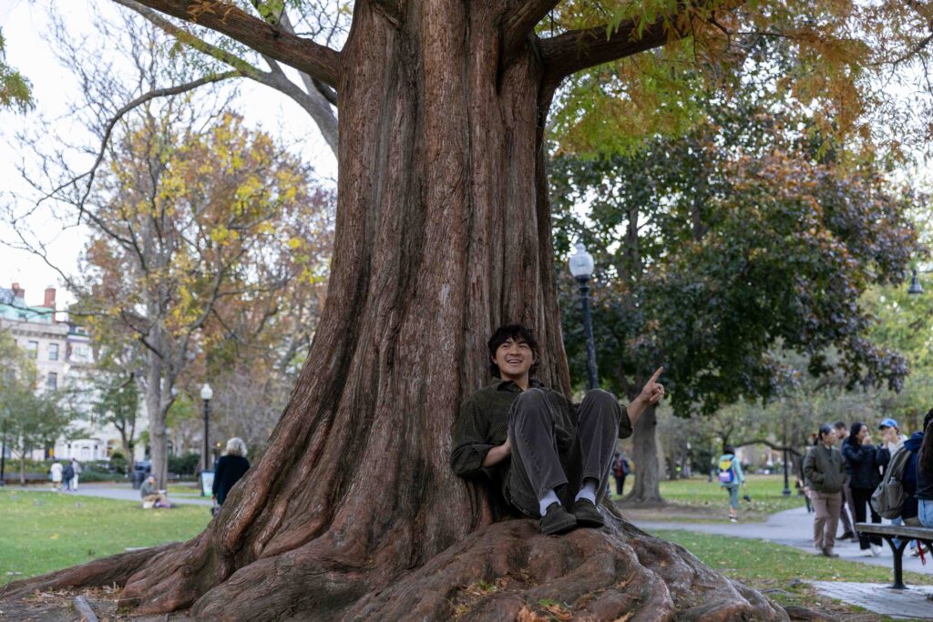 A man sits smiling while sitting at the base of a large tree