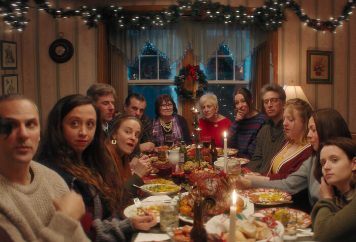 Family gathered around table covered in dishes, candles, with Christmas decorations on wall and window behind them