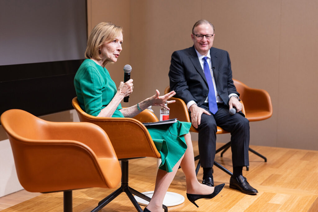 Judy Woodruff, in green dress, sits and speaks into a mic as Jay Bernhardt, in blue suit, looks on