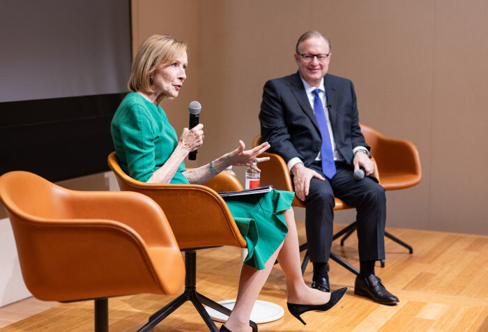 Judy Woodruff, in green dress, sits and speaks into a mic as Jay Bernhardt, in blue suit, looks on