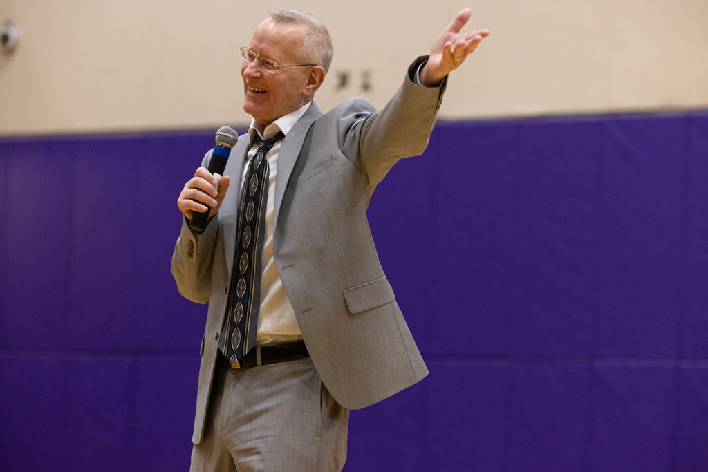 White haired man in tan suit, tie smiles, gestures into a microphone