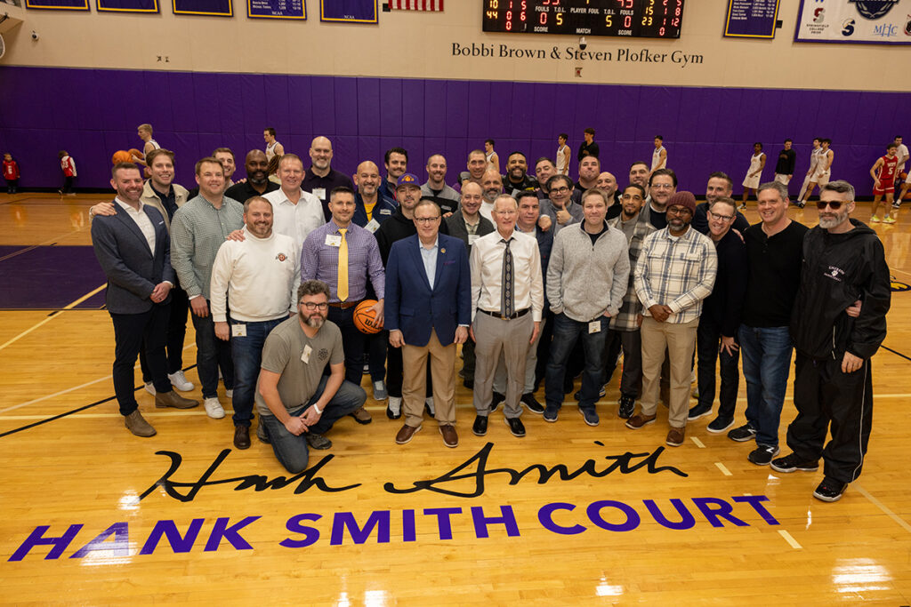 Group of men pose on a basketball court behind the words Hank Smith Court