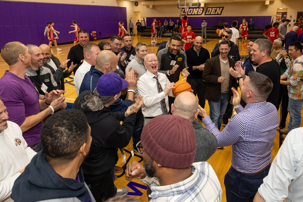 Man with white hair holding a basketball laughs as men stand around him in a circle clapping