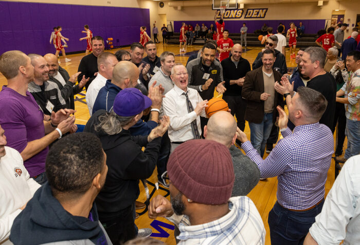 Man with white hair holding a basketball laughs as men stand around him in a circle clapping