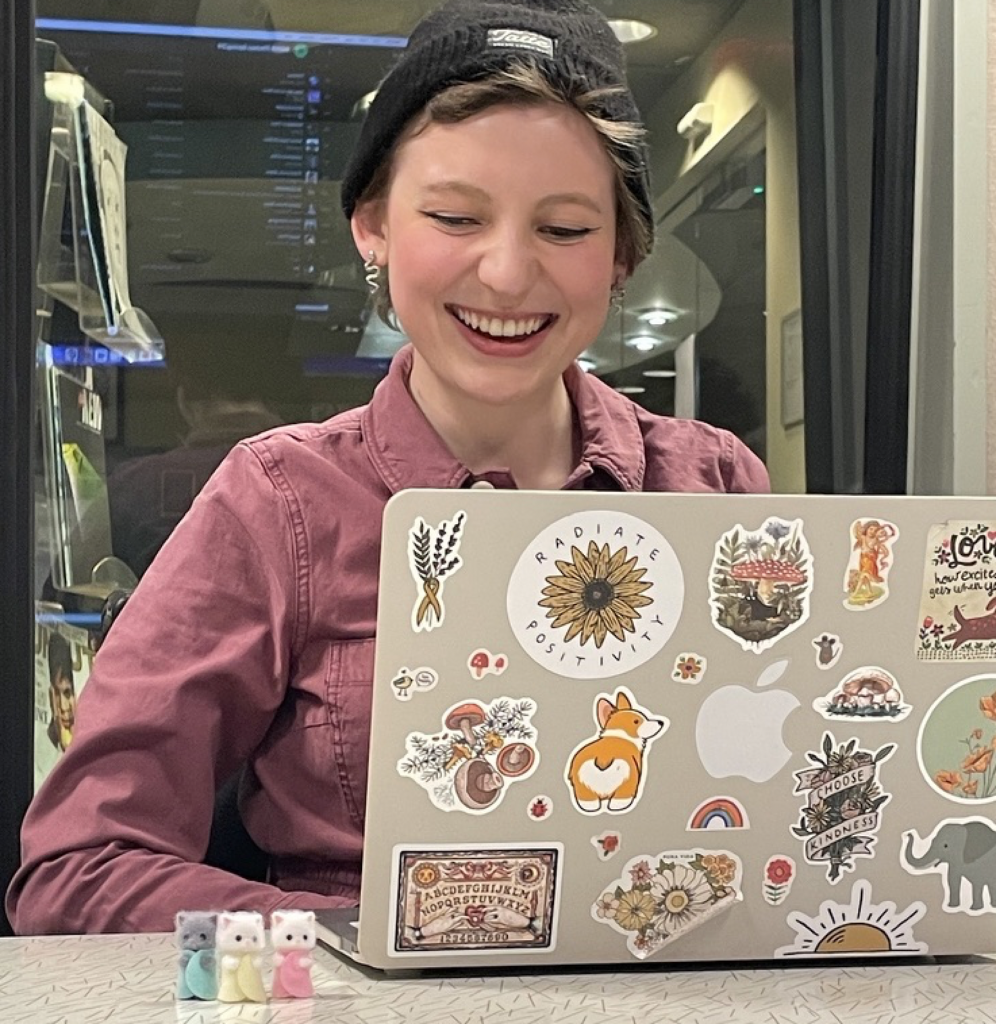 Nora Onanian smiling in front of her laptop, with three little Calico Critters toys on the table in front of her.