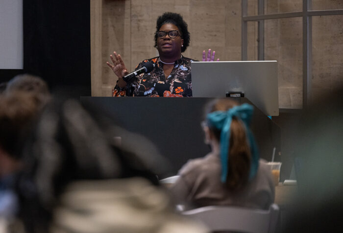 Woman in floral shirt stands behind podium, gesturing