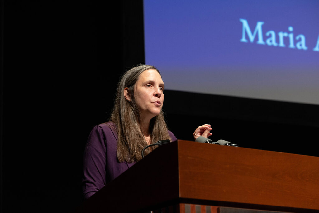 Woman in purple jacket with long brown hair speaks at podium